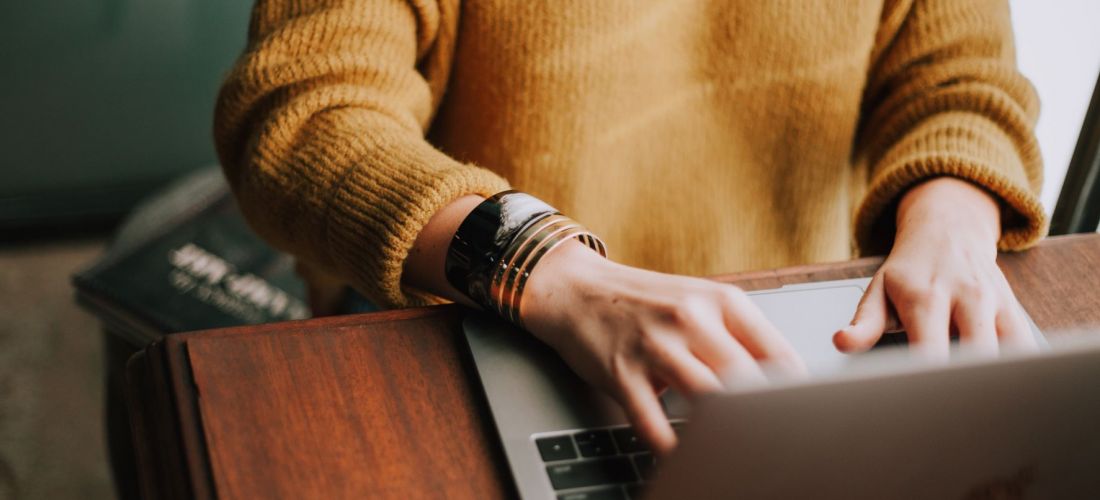 Foto einer Person mit senfgelben Pullover an einem Holztisch sitzend und auf einem Laptop schreibend