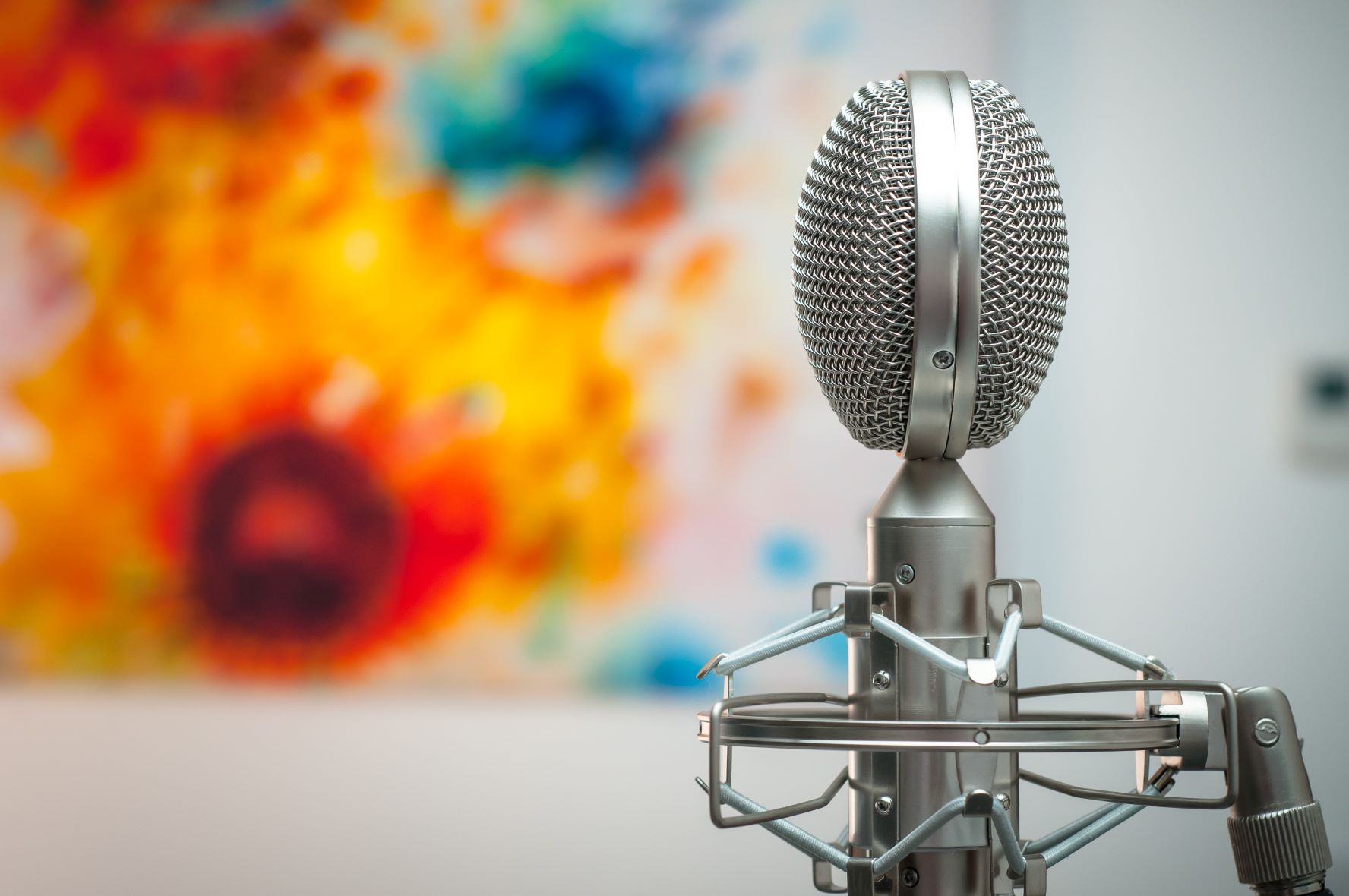 Das Bild zeit ein silbernes Mikrofon, wie es etwa für Radio- oder Podcastaufzeichnungen benutzt wird. Im Hintergrund ist verschwommen ein abstraktes Bild mit kräftigen Gelb- und Rottönen.